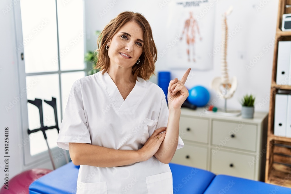 Fototapeta premium Middle age physiotherapist woman working at pain recovery clinic with a big smile on face, pointing with hand and finger to the side looking at the camera.