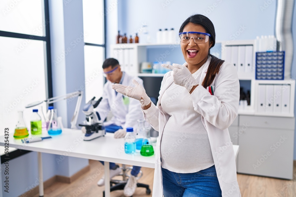 Young hispanic woman expecting a baby working at scientist laboratory amazed and smiling to the camera while presenting with hand and pointing with finger. Young hispanic woman expecting a baby working at scientist laboratory amazed and smiling to the camera while presenting with hand and pointing with finger.