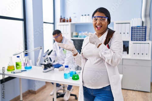 Young hispanic woman expecting a baby working at scientist laboratory amazed and smiling to the camera while presenting with hand and pointing with finger.