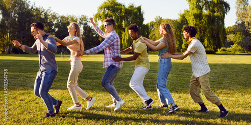 Cheerful active young men and women having fun running in row one after another during gathering in park. Funny multiracial friends have fun together outdoors on summer day. Web banner.