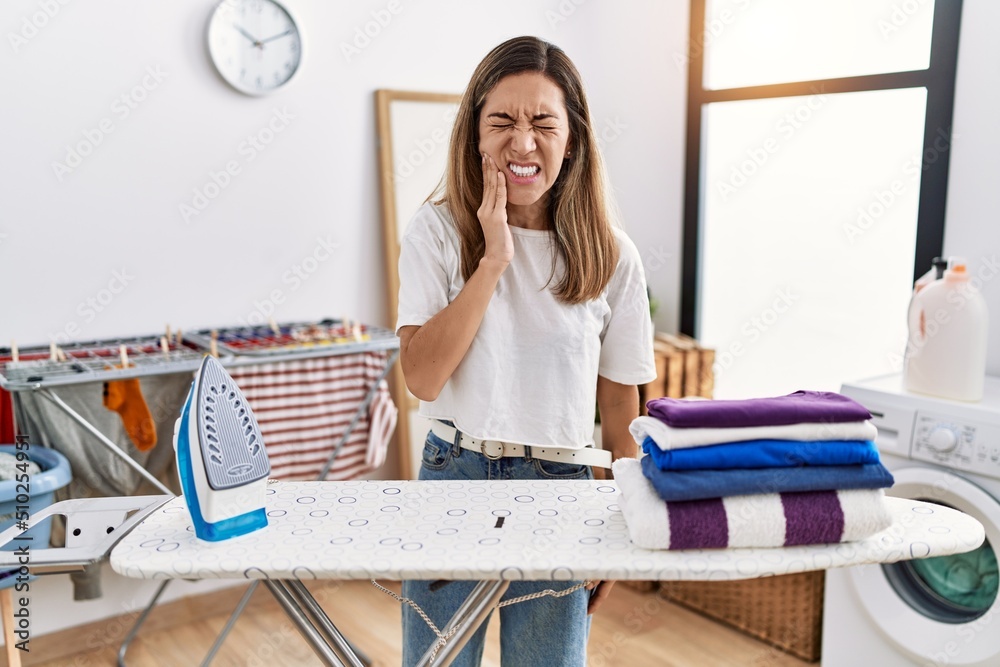 Young hispanic woman ironing clothes at laundry room touching mouth with hand with painful expression because of toothache or dental illness on teeth. dentist Young hispanic woman ironing clothes at laundry room touching mouth with hand with painful expression because of toothache or dental illness on teeth. dentist