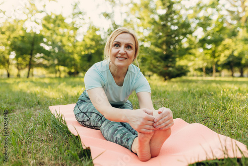 Middle age woman relaxing on green grass during yoga exercise, morning routine, sunset light, outdoor. Mature female in blue sportswear doing stretching. Age, healthy lifestyle.
