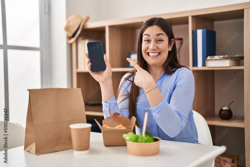 Young brunette woman eating take away food at home showing smartphone screen smiling happy pointing with hand and finger Young brunette woman eating take away food at home showing smartphone screen smiling happy pointing with hand and finger