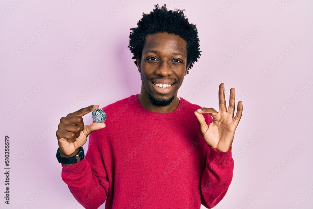 Young african american man holding virtual currency ethereum coin doing ok sign with fingers, smiling friendly gesturing excellent symbol Young african american man holding virtual currency ethereum coin doing ok sign with fingers, smiling friendly gesturing excellent symbol