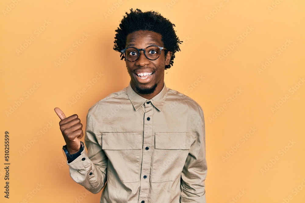Young african american man wearing casual clothes and glasses smiling with happy face looking and pointing to the side with thumb up. Young african american man wearing casual clothes and glasses smiling with happy face looking and pointing to the side with thumb up.