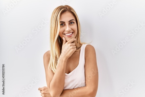Blonde beautiful young woman standing over white isolated background looking confident at the camera smiling with crossed arms and hand raised on chin. thinking positive.
