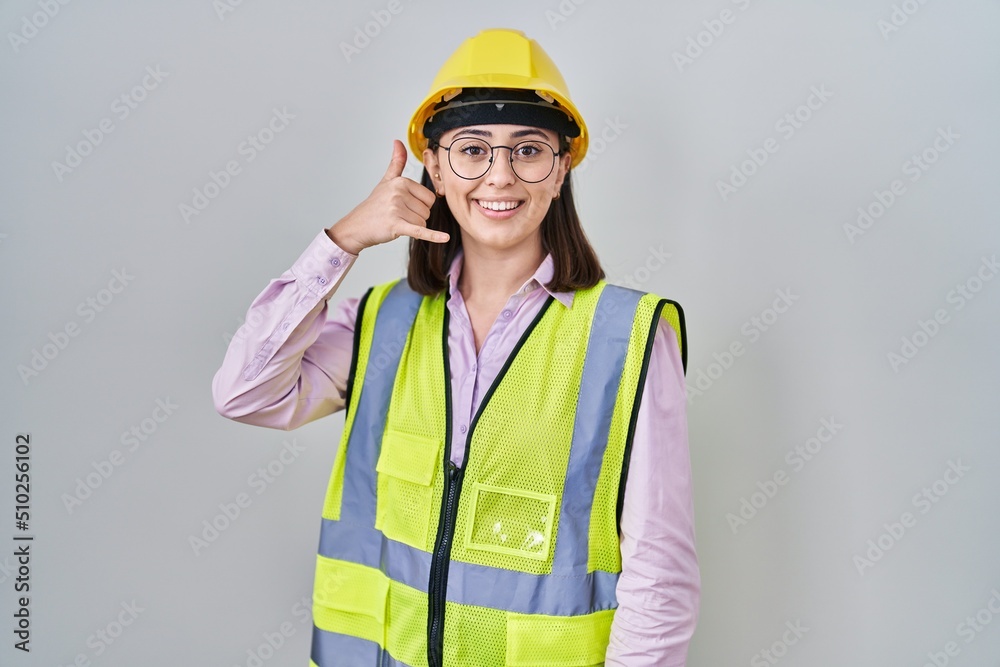 Hispanic girl wearing builder uniform and hardhat smiling doing phone gesture with hand and fingers like talking on the telephone. communicating concepts. Hispanic girl wearing builder uniform and hardhat smiling doing phone gesture with hand and fingers like talking on the telephone. communicating concepts.