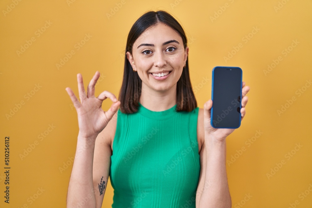 Hispanic girl holding smartphone showing screen doing ok sign with fingers, smiling friendly gesturing excellent symbol Hispanic girl holding smartphone showing screen doing ok sign with fingers, smiling friendly gesturing excellent symbol