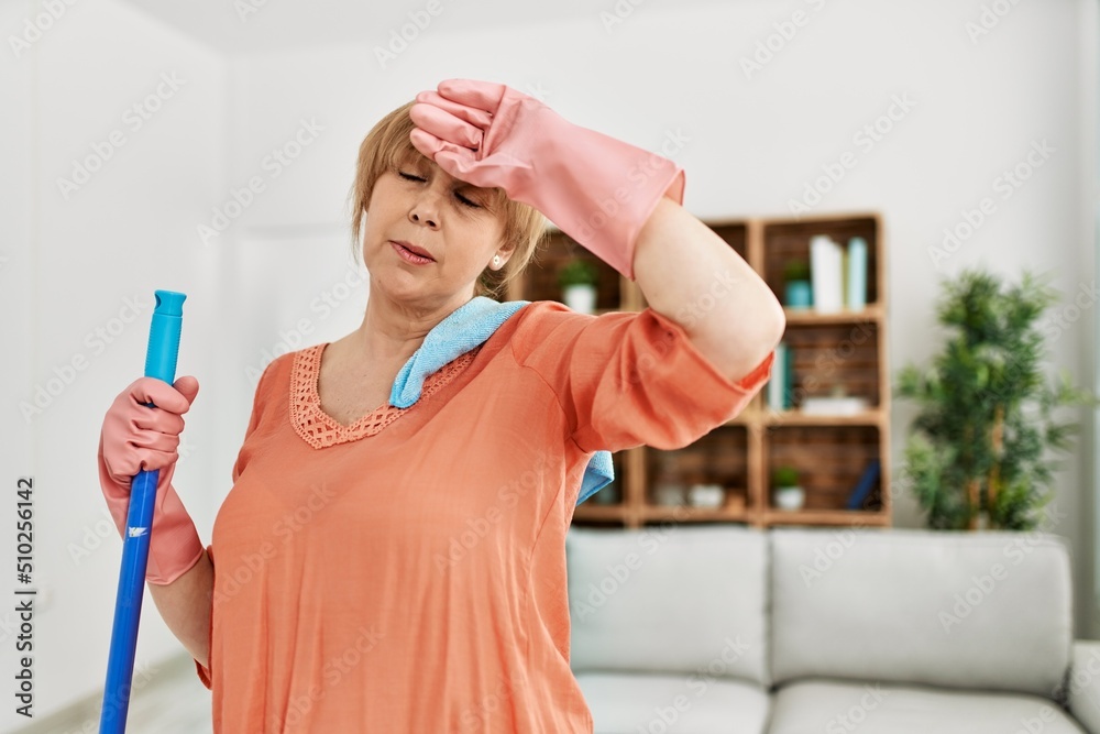 Middle age cleaner woman exhausted with hand on head standing at home. Middle age cleaner woman exhausted with hand on head standing at home.