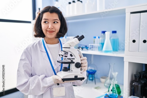Young hispanic woman working at scientist laboratory holding microscope smiling looking to the side and staring away thinking.