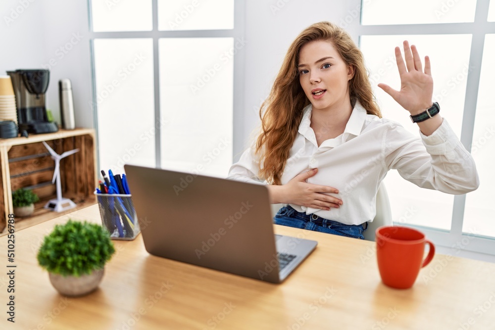 Young caucasian woman working at the office using computer laptop swearing with hand on chest and open palm, making a loyalty promise oath Young caucasian woman working at the office using computer laptop swearing with hand on chest and open palm, making a loyalty promise oath