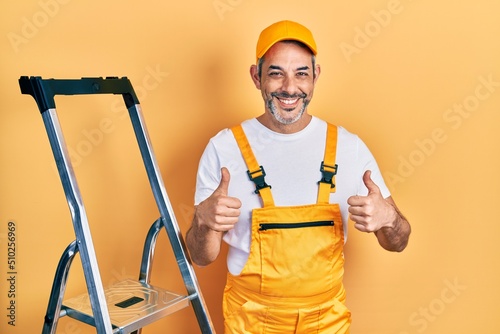 Handsome middle age man with grey hair holding ladder success sign doing positive gesture with hand, thumbs up smiling and happy. cheerful expression and winner gesture. photo