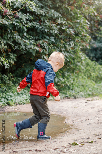 Child in a red waterproof coat in the rain jumping in puddles. A boy stands with his head bowed, a bright raincoat. Kid playing autumn park. Outdoor fun by any weather. © Linas T