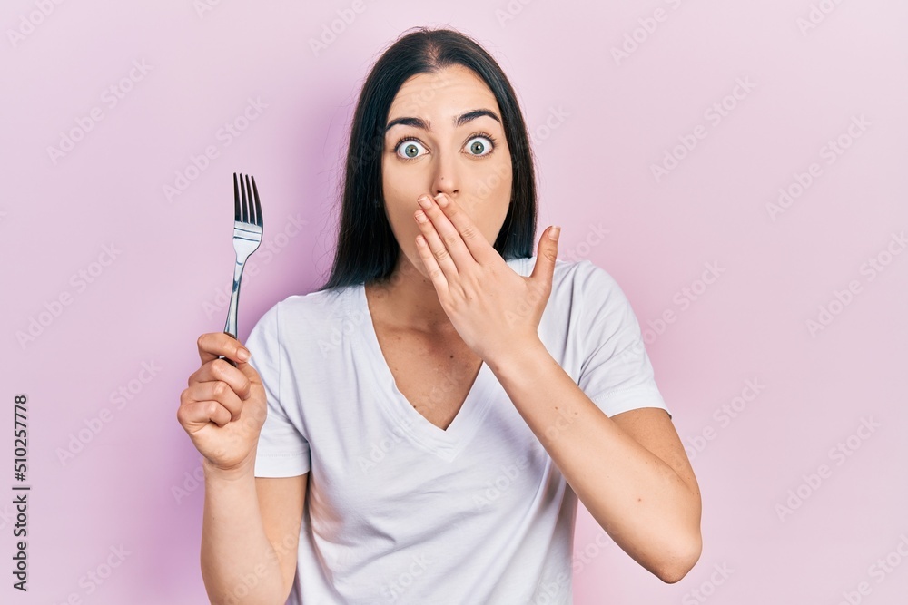 Beautiful woman with blue eyes holding one silver fork covering mouth with hand, shocked and afraid for mistake. surprised expression Beautiful woman with blue eyes holding one silver fork covering mouth with hand, shocked and afraid for mistake. surprised expression