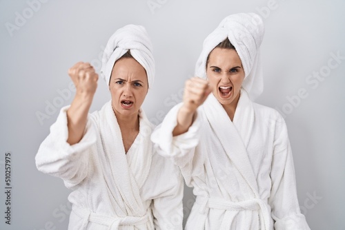 Middle age woman and daughter wearing white bathrobe and towel angry and mad raising fist frustrated and furious while shouting with anger. rage and aggressive concept.