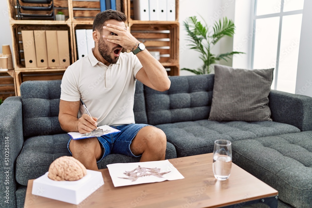 Handsome hispanic man working with rorschach test at psychology clinic peeking in shock covering face and eyes with hand, looking through fingers with embarrassed expression.