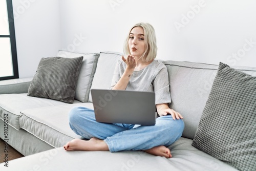 Young caucasian woman using laptop at home sitting on the sofa looking at the camera blowing a kiss with hand on air being lovely and sexy. love expression.
