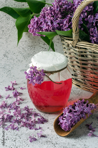 Lilac flowers syrup in glass jar and basket with flowers on grey background.