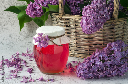 Healthy syrup with lilac flowers in glass jar and basket with flowers on grey background.