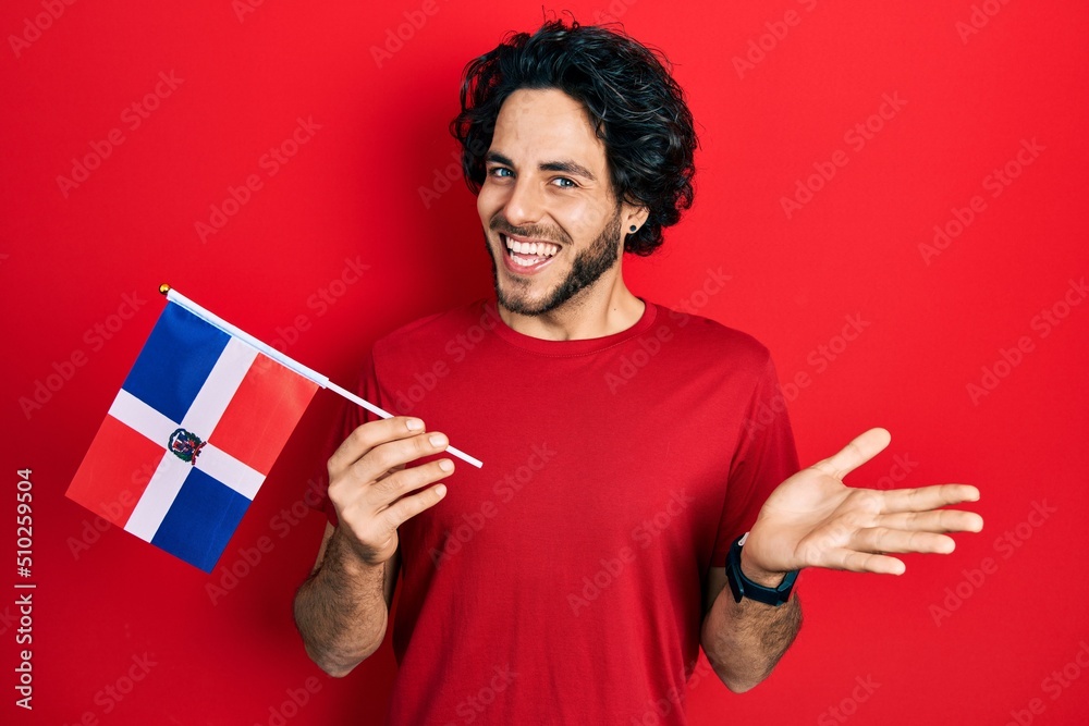 Handsome hispanic man holding dominican republic flag celebrating achievement with happy smile and winner expression with raised hand Handsome hispanic man holding dominican republic flag celebrating achievement with happy smile and winner expression with raised hand