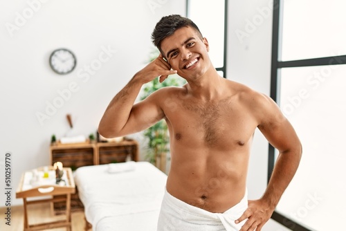 Young hispanic man standing shirtless at spa center smiling doing phone gesture with hand and fingers like talking on the telephone. communicating concepts.