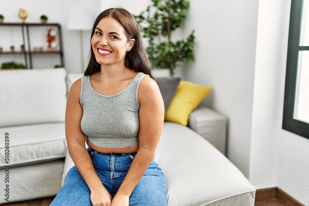 Young brunette woman sitting on the sofa at home looking away to side with smile on face, natural expression. laughing confident. Young brunette woman sitting on the sofa at home looking away to side with smile on face, natural expression. laughing confident.