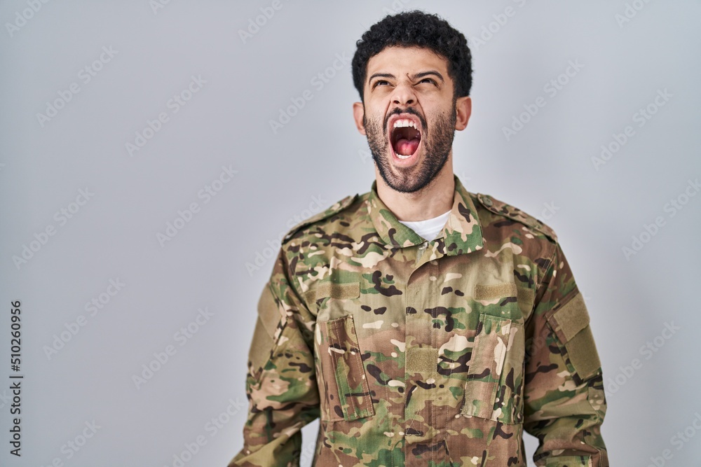 Arab man wearing camouflage army uniform angry and mad screaming frustrated and furious, shouting with anger. rage and aggressive concept. Arab man wearing camouflage army uniform angry and mad screaming frustrated and furious, shouting with anger. rage and aggressive concept.