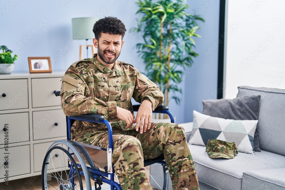 Arab man wearing camouflage army uniform sitting on wheelchair winking looking at the camera with sexy expression, cheerful and happy face.