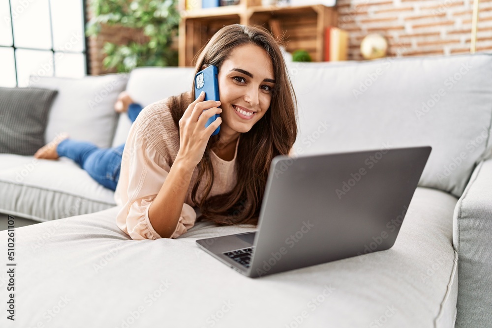 Naklejka premium Young hispanic woman smiling confident using laptop and talking on the smartphone at home