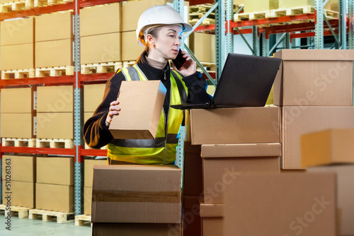 Warehouse worker woman. Warehouse manager with laptop. Girl is talking on phone standing in front of boxes. Warehouse worker near shelving. Career in distribution center. Storekeeper in yellow vest