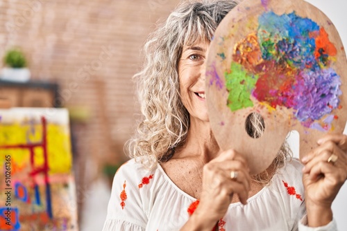 Middle age woman holding painter palette covering face smiling with a happy and cool smile on face. showing teeth.