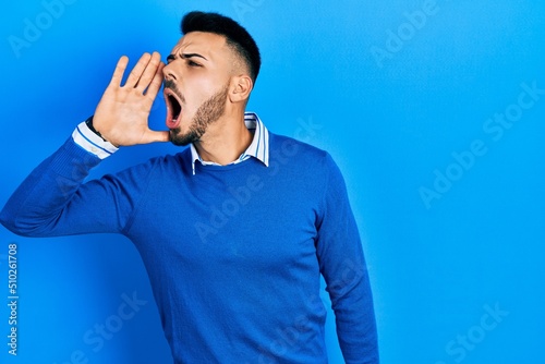 Young hispanic man with beard wearing casual blue sweater shouting and screaming loud to side with hand on mouth. communication concept.