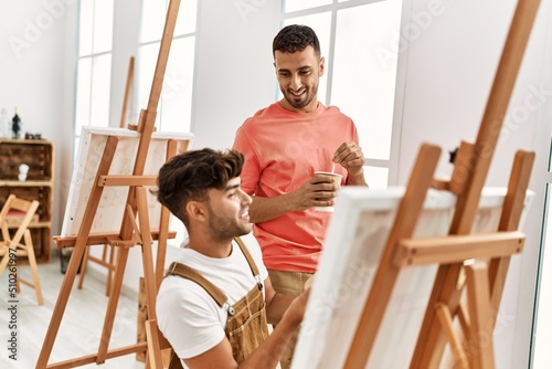 Two hispanic men couple smiling confident drinking coffee and drawing at art studio