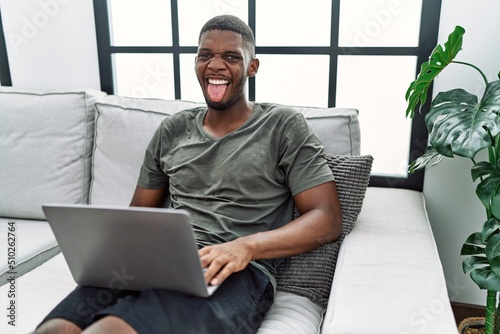 Young african american man using laptop at home sitting on the sofa sticking tongue out happy with funny expression. emotion concept.