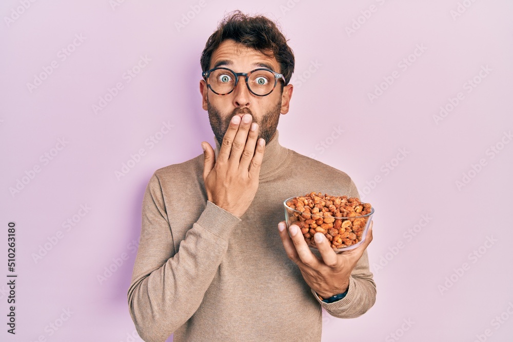 Handsome man with beard holding peanuts covering mouth with hand, shocked and afraid for mistake. surprised expression Handsome man with beard holding peanuts covering mouth with hand, shocked and afraid for mistake. surprised expression
