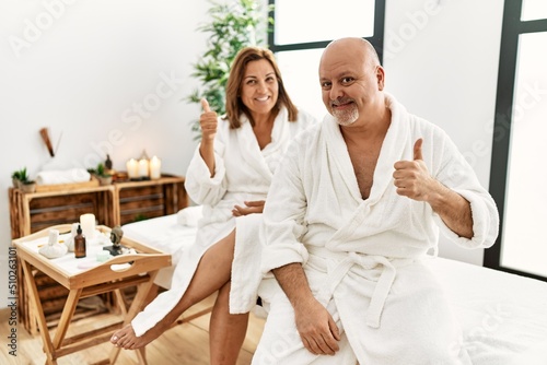 Middle age hispanic couple wearing bathrobe at wellness spa doing happy thumbs up gesture with hand. approving expression looking at the camera showing success.