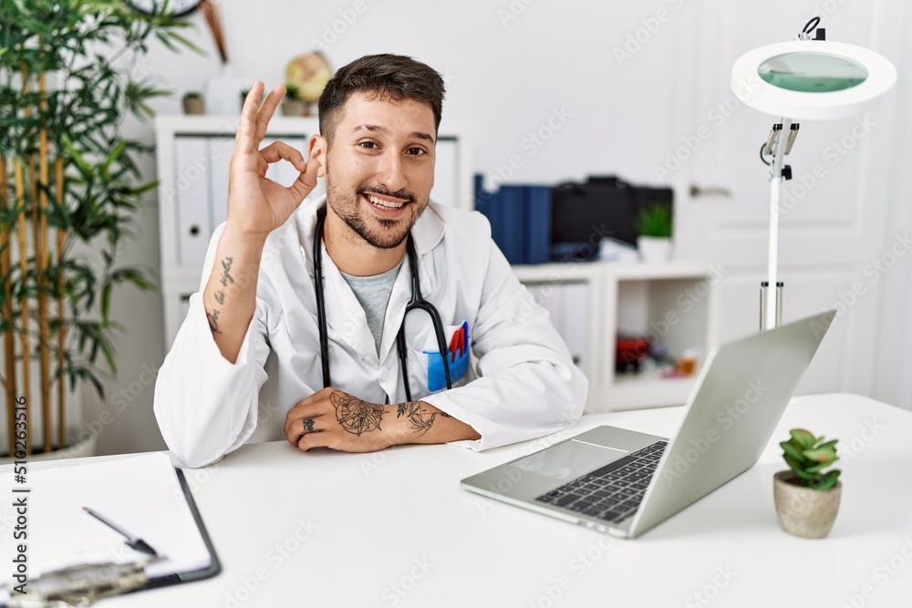 Young doctor working at the clinic using computer laptop smiling positive doing ok sign with hand and fingers. successful expression. Young doctor working at the clinic using computer laptop smiling positive doing ok sign with hand and fingers. successful expression.