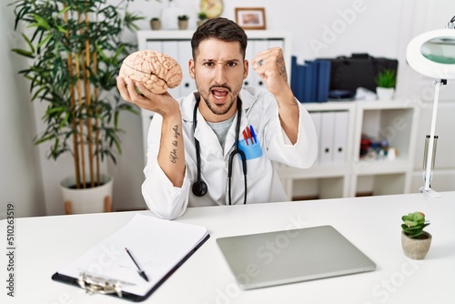 Young doctor holding brain at medical clinic annoyed and frustrated shouting with anger, yelling crazy with anger and hand raised