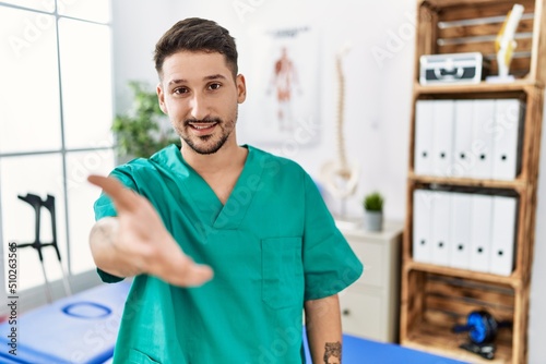 Young physiotherapist man working at pain recovery clinic smiling cheerful offering palm hand giving assistance and acceptance.