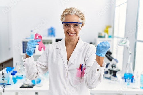 Middle age blonde woman working at laboratory looking for breast cancer cure screaming proud, celebrating victory and success very excited with raised arms