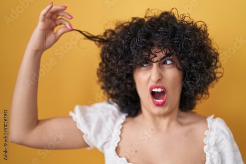 Young brunette woman with curly hair holding curl angry and mad screaming frustrated and furious  shouting with anger. rage and aggressive concept.