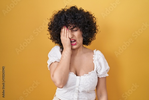 Young brunette woman with curly hair standing over yellow background yawning tired covering half face, eye and mouth with hand. face hurts in pain.