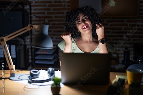 Young brunette woman with curly hair working at the office at night very happy and excited doing winner gesture with arms raised, smiling and screaming for success. celebration concept.