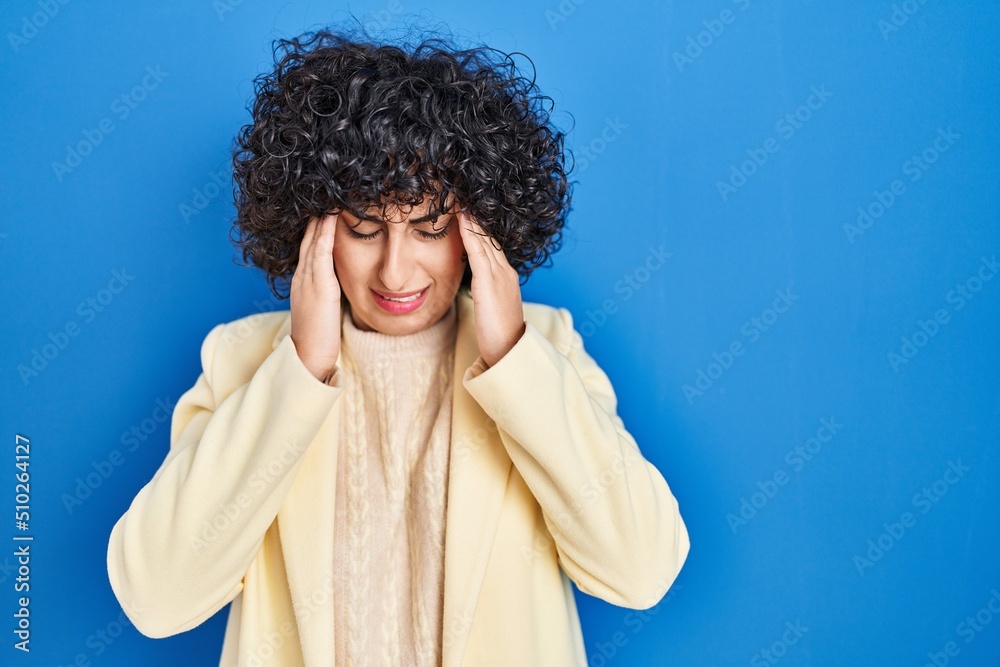 Young brunette woman with curly hair standing over blue background with hand on head, headache because stress. suffering migraine. Young brunette woman with curly hair standing over blue background with hand on head, headache because stress. suffering migraine.