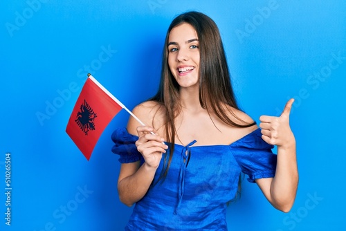 Young brunette teenager holding albania flag smiling happy and positive, thumb up doing excellent and approval sign photo