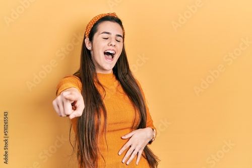 Young brunette teenager wearing casual yellow sweater laughing at you, pointing finger to the camera with hand over body, shame expression