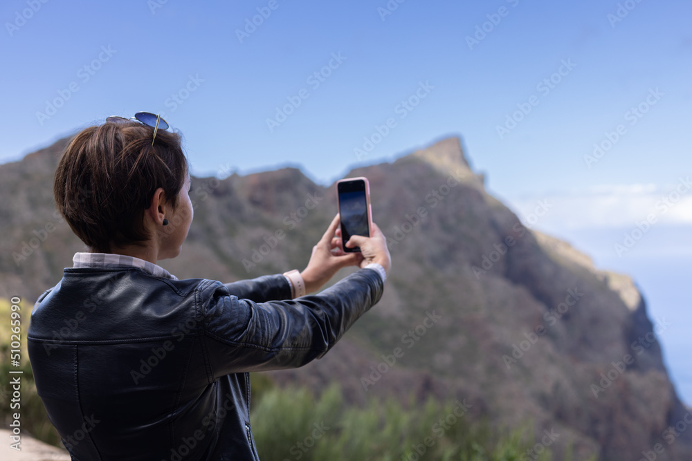 Girl in leather jacket snapping a photo of mountains. Rear view.