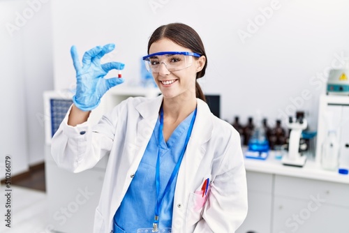 Young hispanic woman wearing scientist uniform holding pill at laboratory
