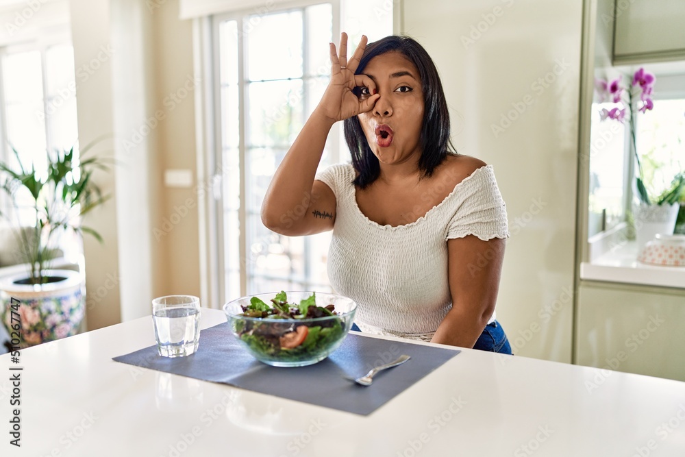 Young hispanic woman eating healthy salad at home doing ok gesture shocked with surprised face, eye looking through fingers. unbelieving expression.