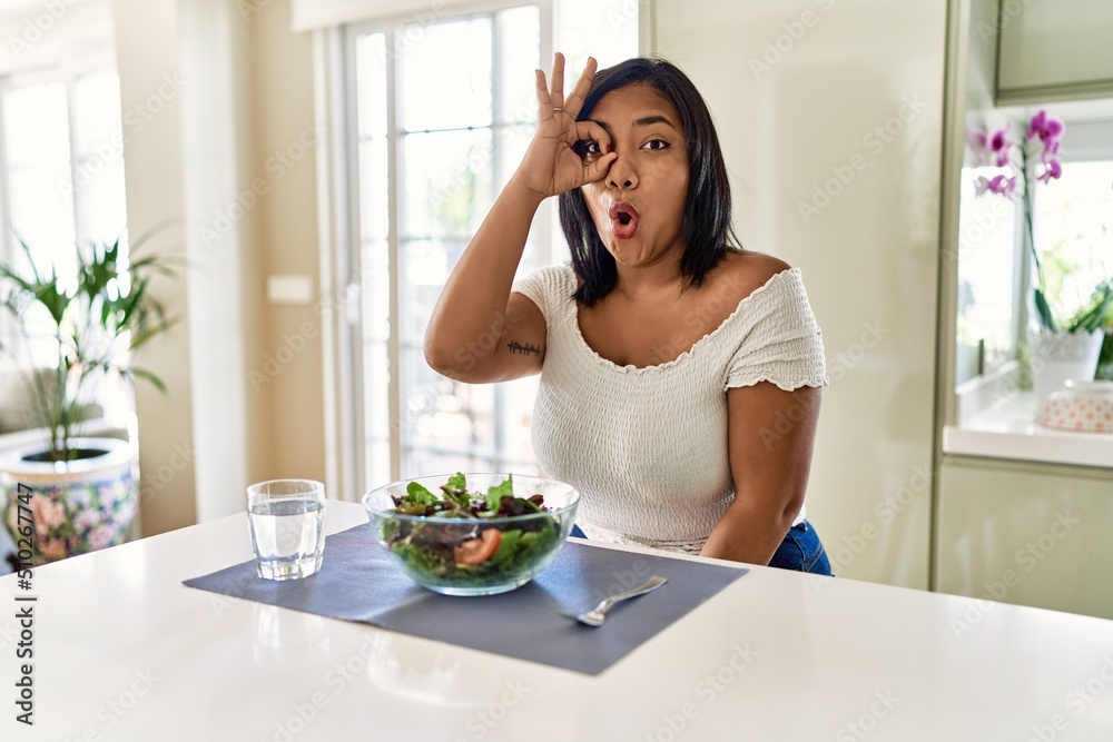 Young hispanic woman eating healthy salad at home doing ok gesture shocked with surprised face, eye looking through fingers. unbelieving expression. Young hispanic woman eating healthy salad at home doing ok gesture shocked with surprised face, eye looking through fingers. unbelieving expression.